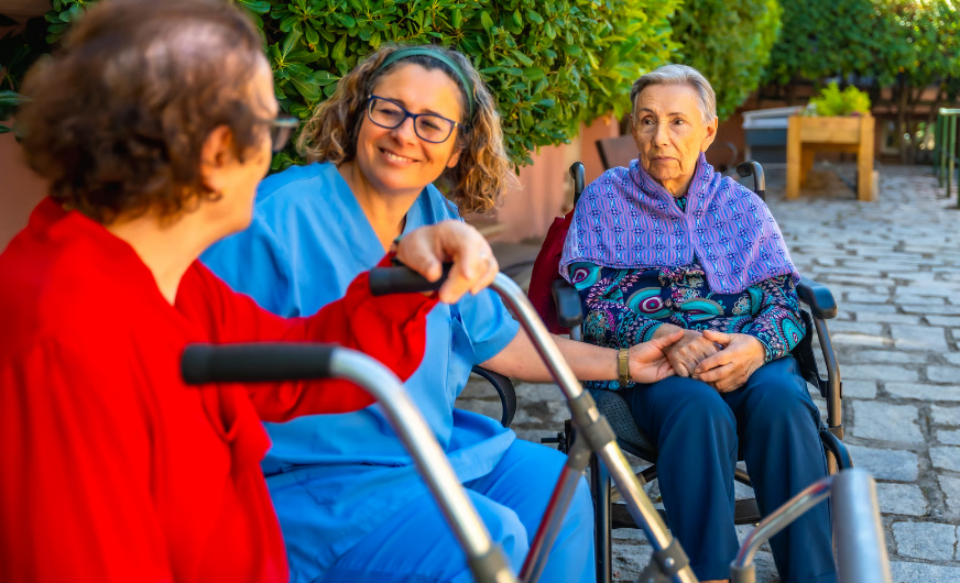 A nurse in blue scrubs and glasses smiles while holding the hand of an elderly woman in a wheelchair. She is also supporting another senior woman in a red shirt who is using a walker. They are in an outdoor courtyard with stone paving and green bushes.