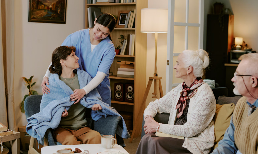 A young female nurse in blue scrubs gently places a blue blanket around a seated woman's shoulders while an elderly couple watches supportively in a cozy living room.
