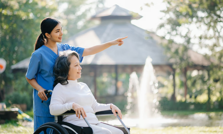 A smiling young nurse in blue scrubs points toward the horizon while standing behind an elderly woman in a wheelchair in a sunny park with a fountain.
