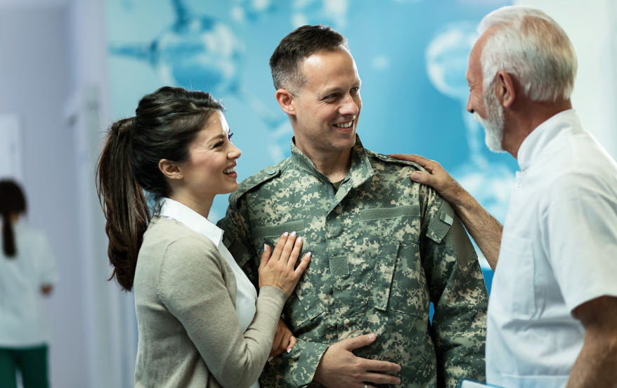 A smiling man in a military camouflage uniform stands with his wife while talking to a senior male doctor in a white medical shirt. The woman has her hand on the soldier's arm, and they are all looking at each other in a brightly lit clinic hallway with a blue molecular graphic on the wall.