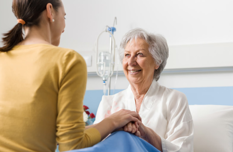 A senior woman in a hospital bed smiling while holding hands with a young woman in a yellow shirt.