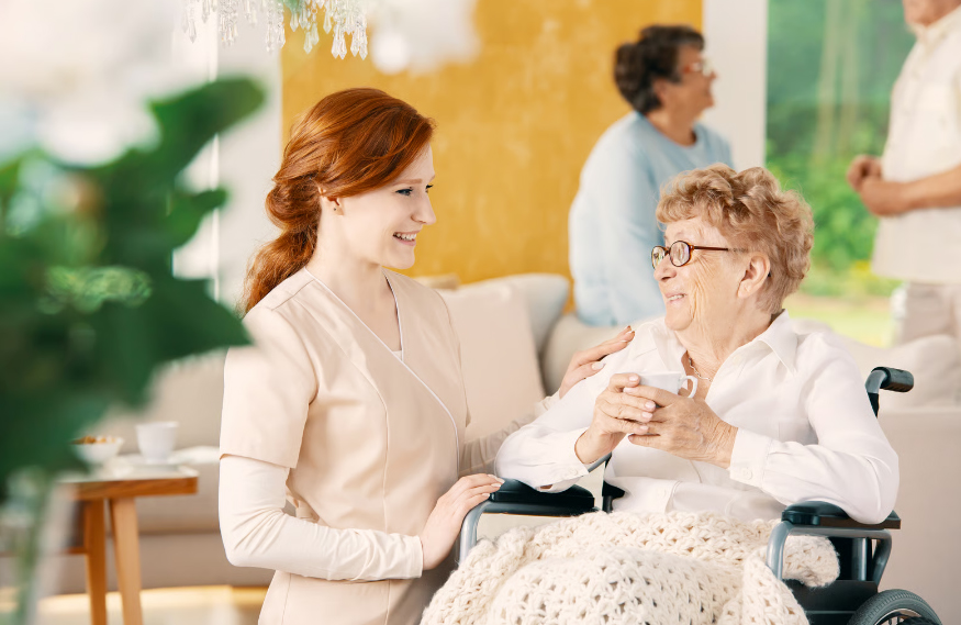 A smiling hospice nurse in a tan uniform standing next to a senior woman in a wheelchair holding a cup.