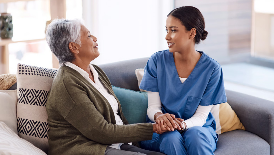 A hospice nurse in blue scrubs sits on a sofa, warmly holding the hands of a senior woman during a supportive conversation in a home setting.