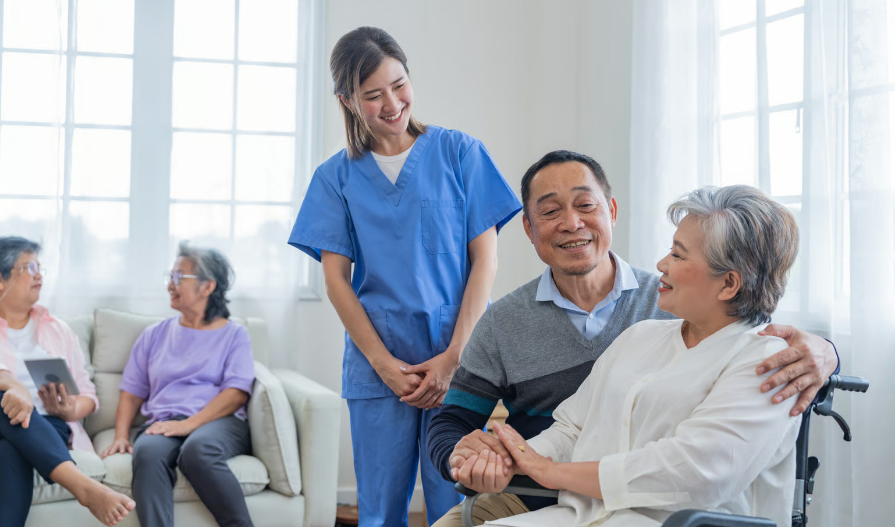 A smiling young female caregiver in blue scrubs standing next to a happy senior East Asian couple holding hands in a bright assisted living facility, with other elderly residents blurred in the background.