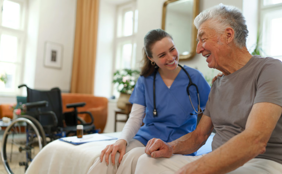 A happy female home care nurse in blue scrubs smiles warmly while talking with a laughing senior man sitting on his bed.