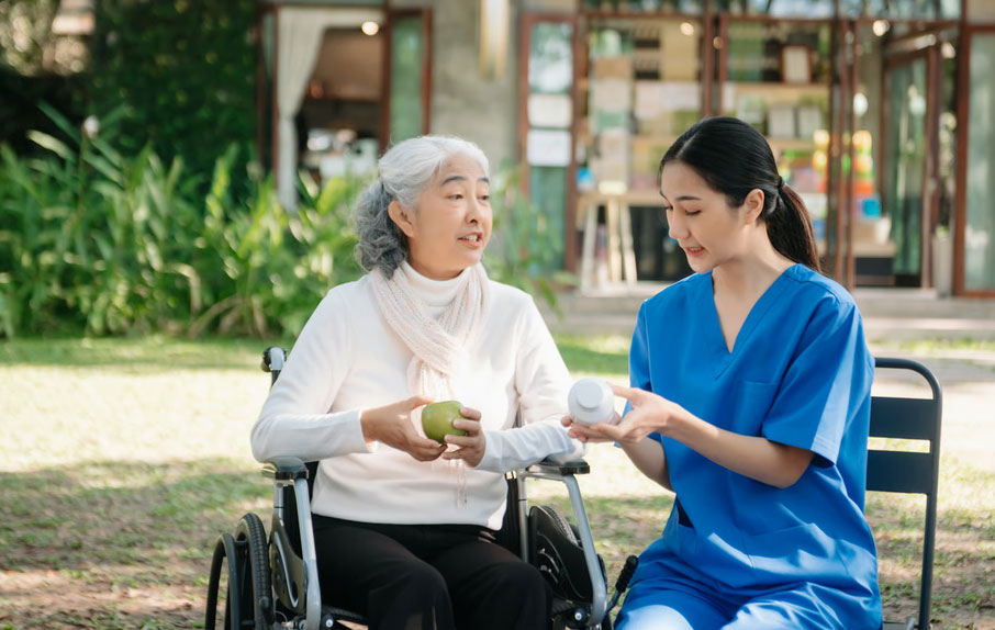 A friendly nurse in blue scrubs reviews medication with an elderly Asian woman in a wheelchair holding a green apple outdoors, showcasing the professional support provided during hospice respite care by iServe Hospice in the Dallas-Fort Worth area.