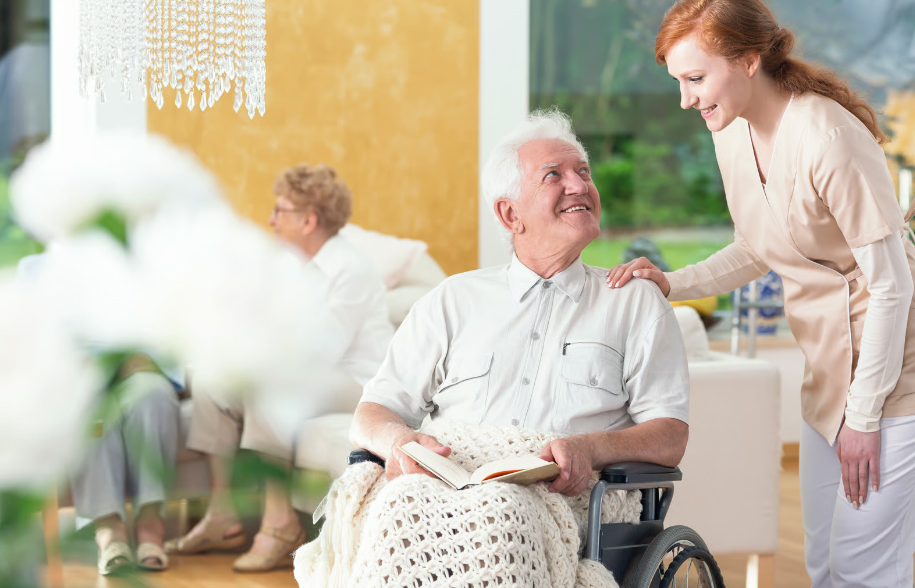 A cheerful young female caregiver in a beige uniform smiling down at an elderly man with white hair sitting in a wheelchair holding a book in a brightly lit, upscale common room.