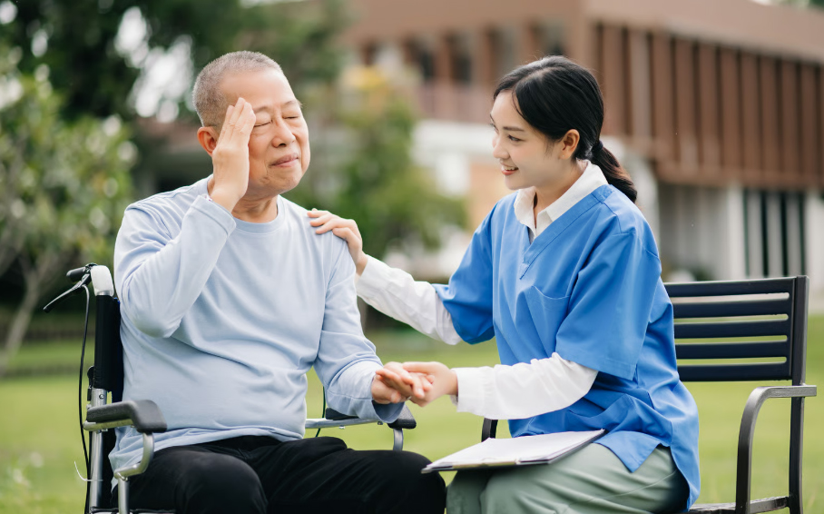 An elderly Asian man in a wheelchair holds his temple as if experiencing a headache or stroke symptom, while a female nurse in blue scrubs offers comfort and support outdoors.