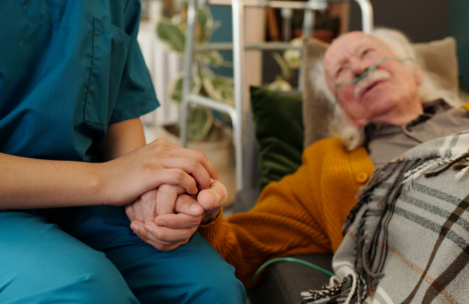 A hospice nurse in blue scrubs holding the hand of an elderly male dementia patient resting in bed with an oxygen tube.