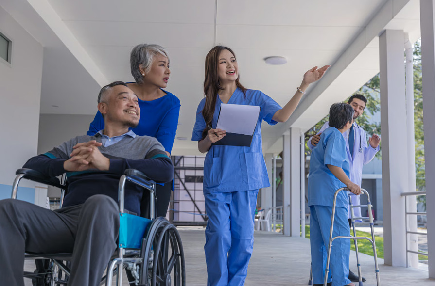 A smiling nurse in blue scrubs holding a clipboard while guiding an elderly man in a wheelchair and his wife down a bright facility hallway.