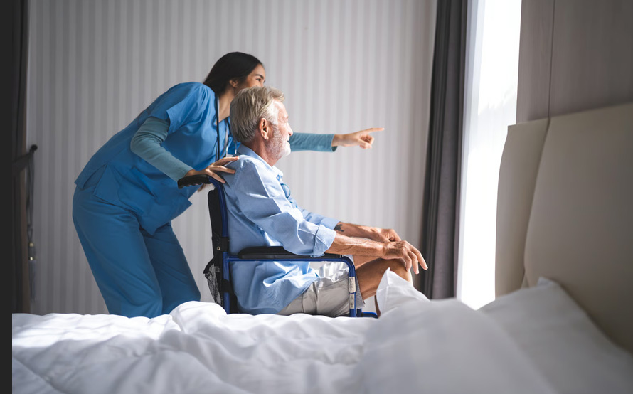 Hospice nurse in blue scrubs assisting an elderly man in a wheelchair as they look out a window, illustrating early hospice enrollment care.