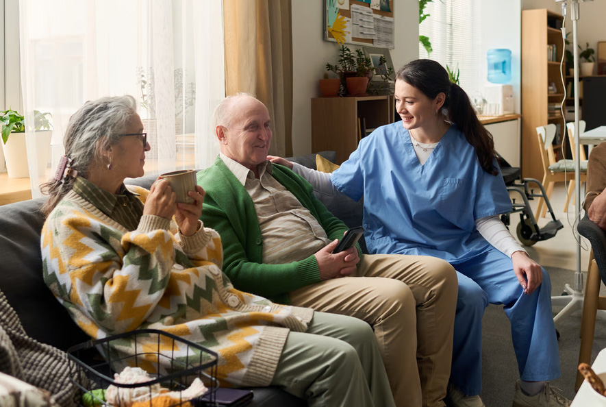 A compassionate hospice nurse in blue scrubs sitting on a couch with an elderly man and woman during a home care consultation for hospice re-election.