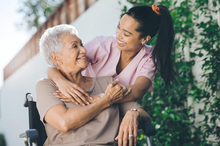 A smiling healthcare caregiver warmly embracing an elderly woman sitting in a wheelchair outdoors, symbolizing health stabilization and positive transition in care.