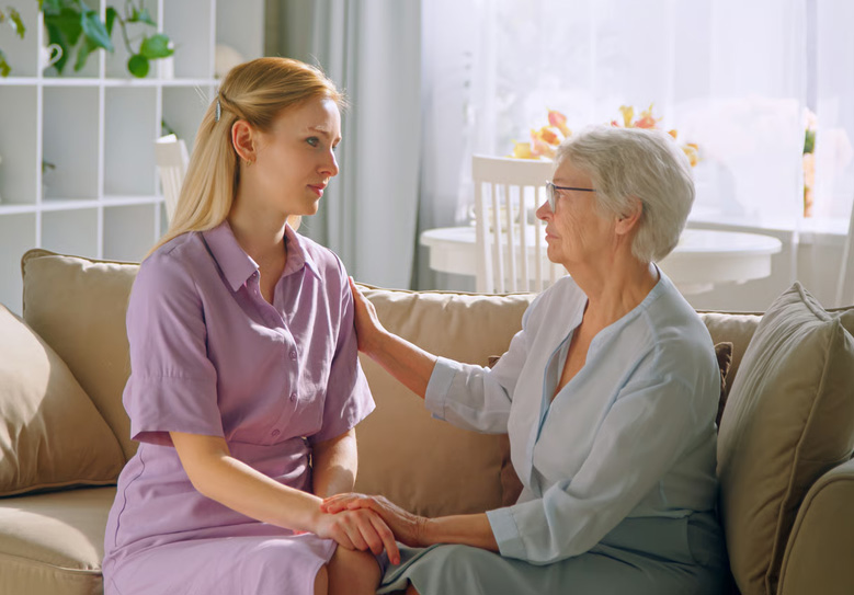 Young female caregiver offering comfort and support to an elderly woman sitting together on a couch at home.