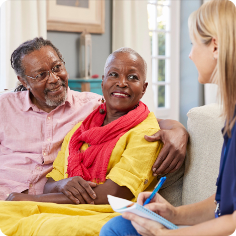 An elderly Black couple sitting on a sofa and smiling warmly while speaking with a hospice care professional who is taking notes.